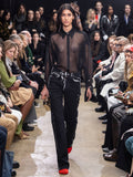 Model Walking in Proenza Schouler Fall Winter 2024 Runway Show Wearing Smithson Shirt in Black Silk Knit, Ryman Jean in Black Garment Printed Denim, Thin Belt in Black Nappa, Crush Clutch in Gold Metal, and Tee Over the Knee Boots in Vermillion Viscose Knit