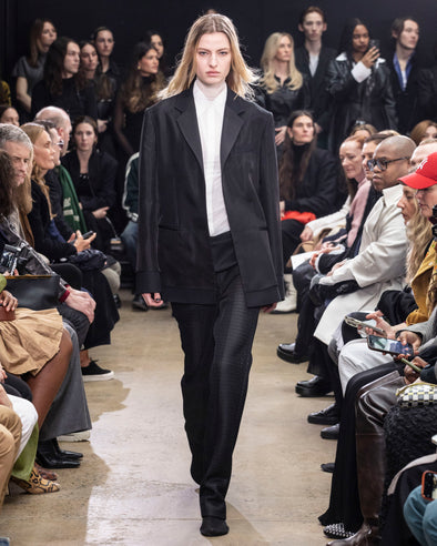 Model Walking in Proenza Schouler Fall Winter 2024 Runway Show Wearing Bronte Jacket in Black Double Weave Viscose Organza, Rio Top in Bone Tulle Jersey, Demi Shirt in Bone Silk Viscose, Chase Pant in Black Technical Mesh Suiting, and Tee Over the Knee Boots in Black Viscose Knit With White Ties