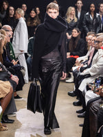 Model Walking in Proenza Schouler Fall Winter 2024 Runway Show Wearing Yara Sweater in Black Double Face Cashmere, Anastasia Top in Black Sheer Crepe Jersey, Adriana Skirt in Black Nappa, Tube Scarf in Black Cashmere Knit, Split Tote in Black Nappa, and  Track Sneakers in Black Nappa/suede