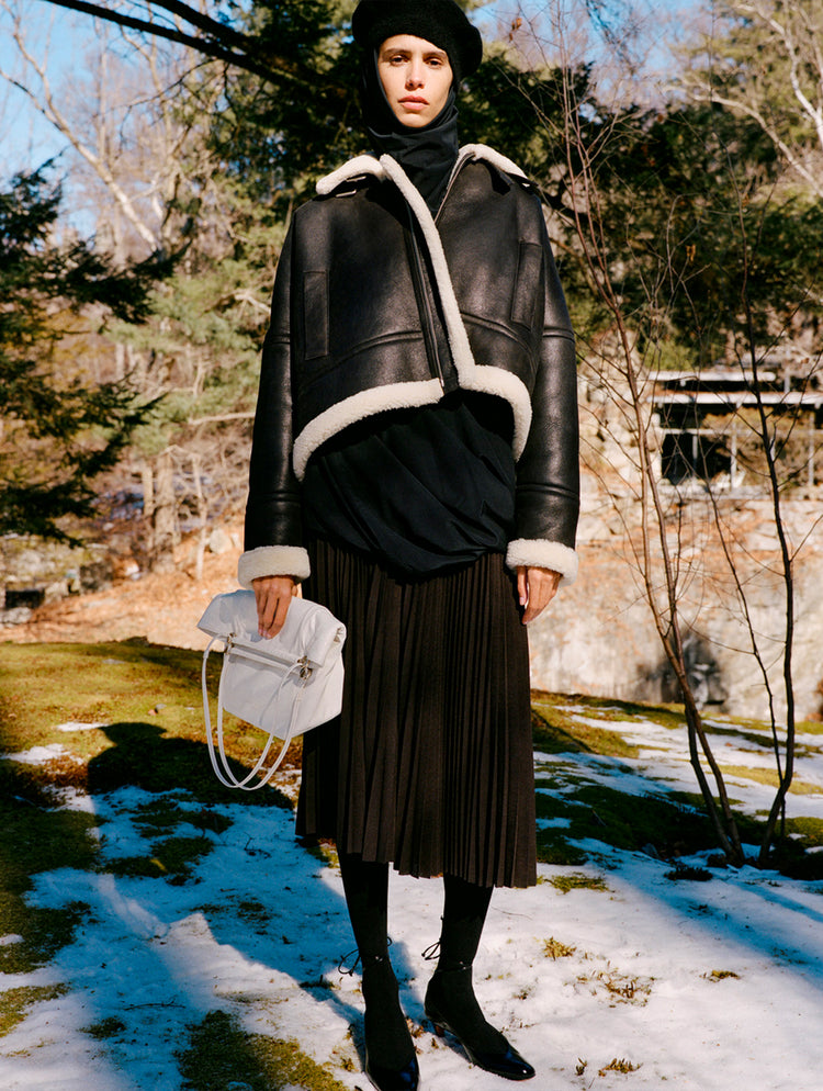 Model standing in snowy field wearing Alya Jacket in Black/Off White Shearling, Solene Top in Anthracite Silk Viscose, Eda Skirt in Espresso Wool Jersey, Beret in Black Wool Knit, Reade Shoulder Bag in Optic White Paper Calf, and Uma Tie Pumps in Black Slick Calf