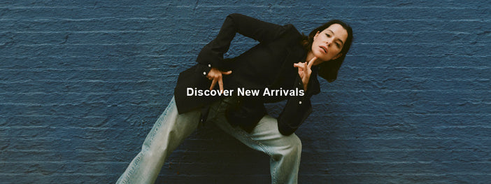 Image of Parker Posey against blue brick wall, wearing Lark Blazer in Wool Gabardine Suiting in black, Ellsworth Jean in classic wash, and Track Sneakers in black/white, 'Discover New Arrivals' overlaid