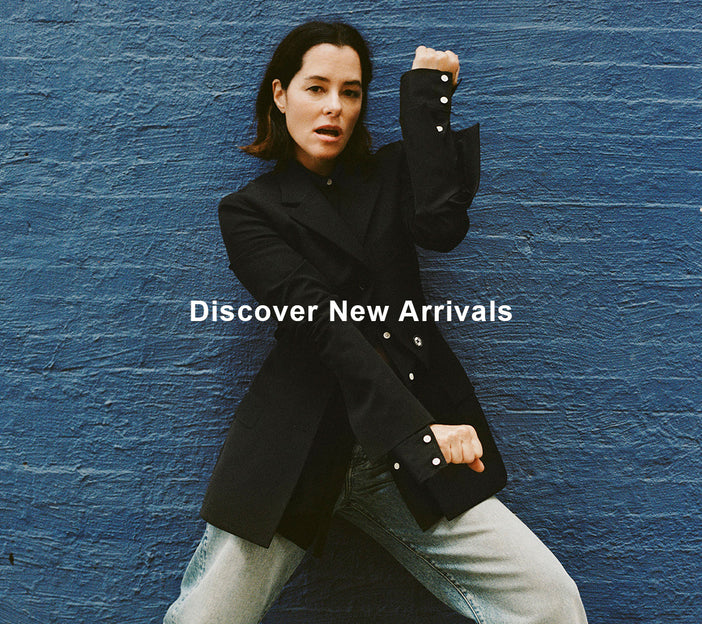 Image of Parker Posey against blue brick wall, wearing Lark Blazer in Wool Gabardine Suiting in black, Ellsworth Jean in classic wash, and Track Sneakers in black/white, 'Discover New Arrivals' overlaid