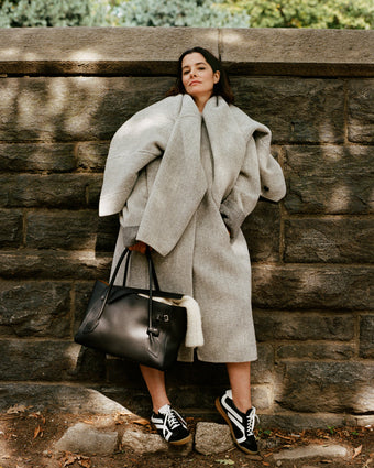 Image of Parker Posey against brick wall, wearing Albers Coat in Textured Double Face Wool in light grey multi and Track Sneakers in black/white, carrying Tate Bag in black