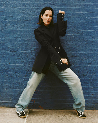 Image of Parker Posey against blue brick wall, wearing Lark Blazer in Wool Gabardine Suiting in black, Ellsworth Jean in classic wash, and Track Sneakers in black/white