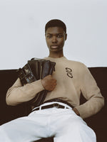 Cropped image of model wearing Harrison Monogram Sweater in Wool Jacquard in oatmeal, Ellsworth Jean in white, carrying City Messenger Bag in black and Flip Shoulder Bag in Eel.