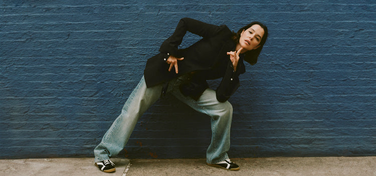 Image of Parker Posey against blue brick wall, wearing Lark Blazer in Wool Gabardine Suiting in black, Ellsworth Jean in classic wash, and Track Sneakers in black/white