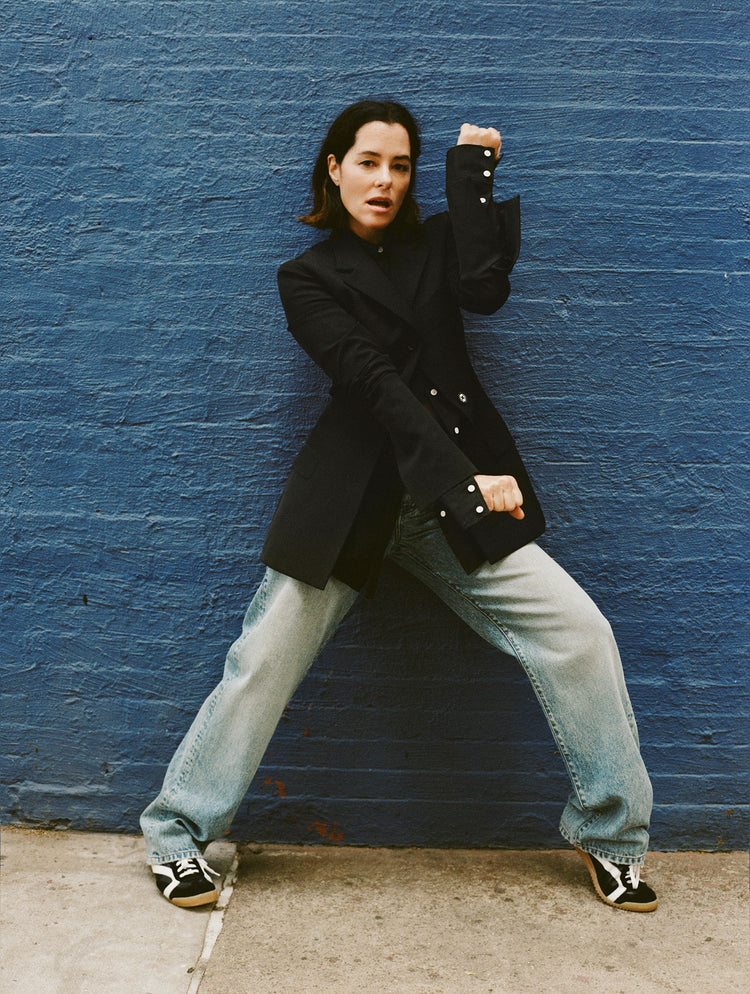 Image of Parker Posey against blue brick wall, wearing Lark Blazer in Wool Gabardine Suiting in black, Ellsworth Jean in classic wash, and Track Sneakers in black/white