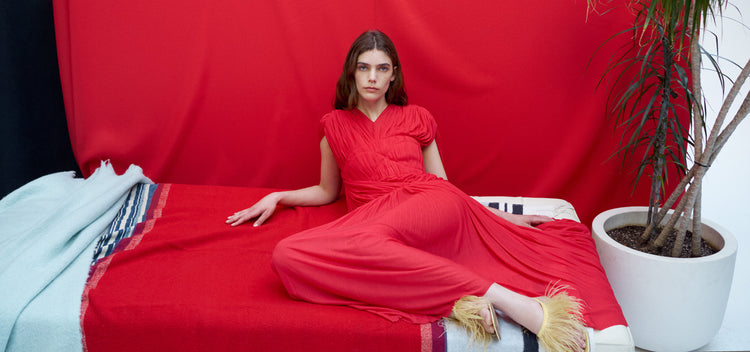 Model laying against tonal red backdrop, wearing Rumi Dress in Pleated Jersey in scarlet and Tee Thong Feather Sandals in resin