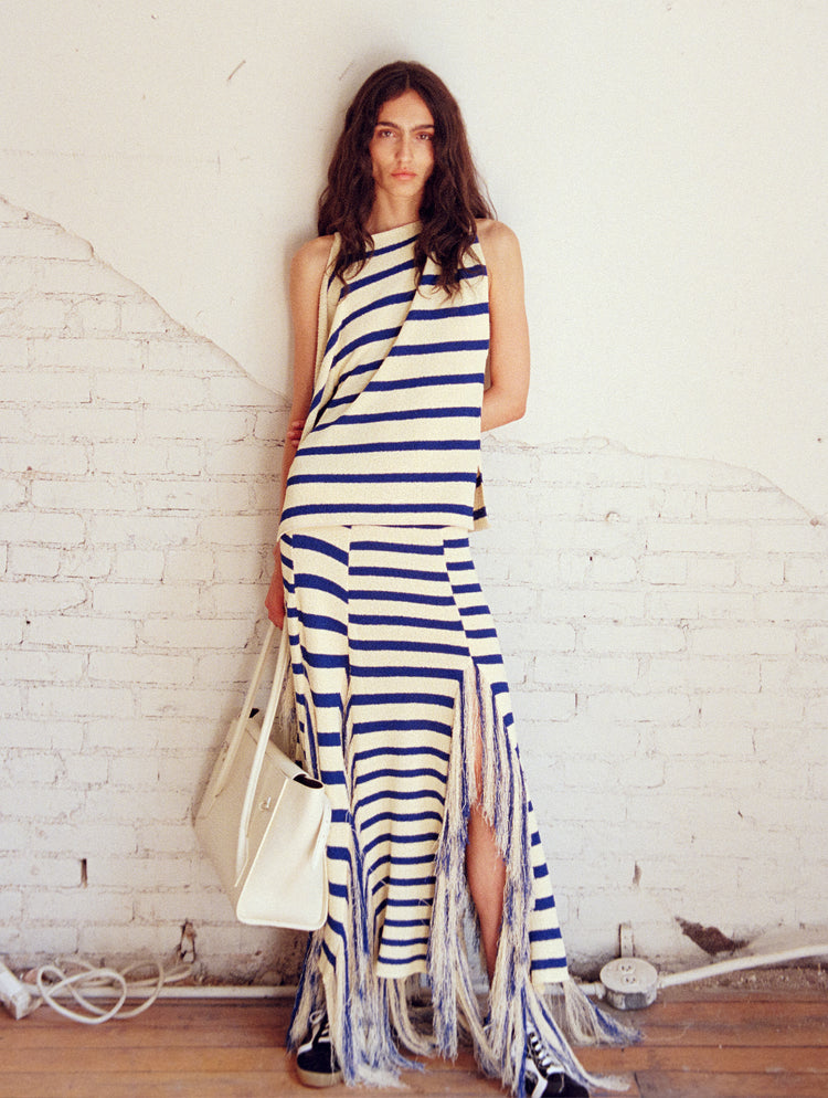 Model standing against white brick wall wearing Yves Top and Skirt in Stripe Fringed Knit in pale yellow multi, holding the Tate Bag in ivory