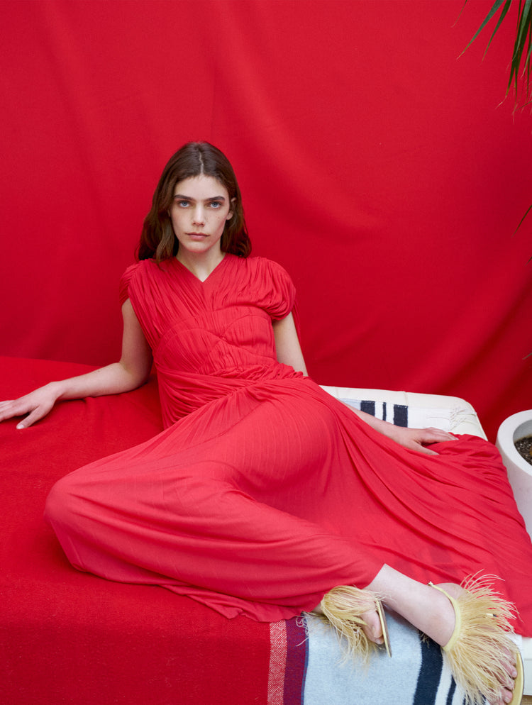 Model laying against tonal red backdrop, wearing Rumi Dress in Pleated Jersey in scarlet and Tee Thong Feather Sandals in resin